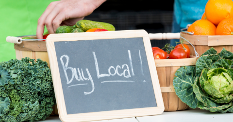 A sign sitting on a table of vegetables reads, "Buy Local!"