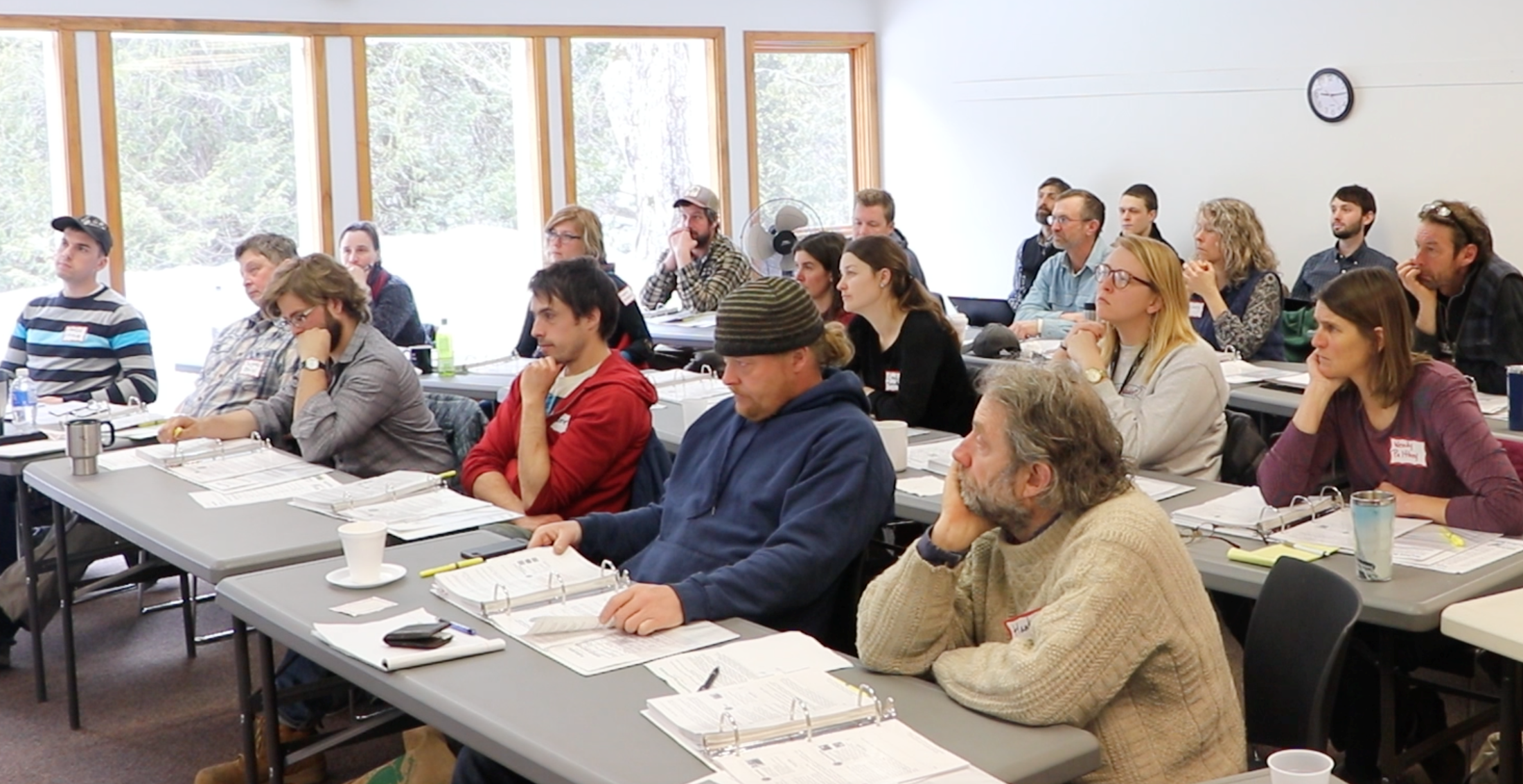 A photo of people sitting in a room at rows of tables facing the front of the room while listening to a presentation.