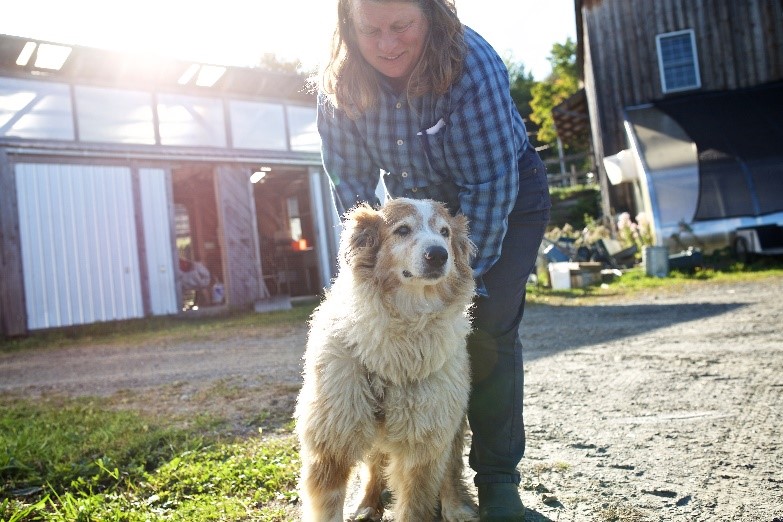 A farmer with a fluffy dog, with farm buildings and equipment in the background