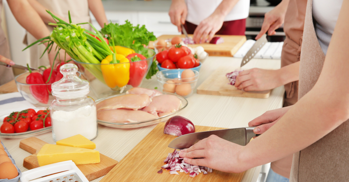 A group of people stand around a table cutting vegetables. There are veggies, meats, and cheeses in the center.