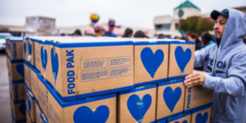 a worker in a sweatshirt and baseball cap stacking cardboard boxes containing food