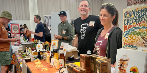 Vendors at a tradeshow.  In the foreground, a table displays cheese products and promotional materials. Two smiling vendors stand behind the table, wearing "Cheese" shirts.