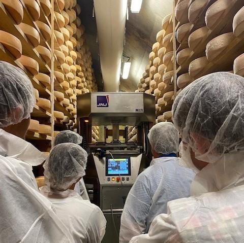 people in white coats and hairnets inside cheese cave with computer device