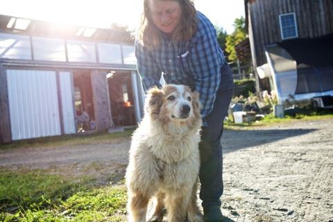A farmer with a fluffy dog, with farm buildings and equipment in the background