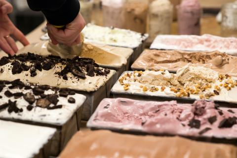 A hand scoops ice cream from one of several metal pans of ice cream