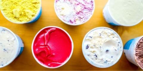 A photo of seven open pint containers of brightly colored ice cream on a light wooden surface viewed from above