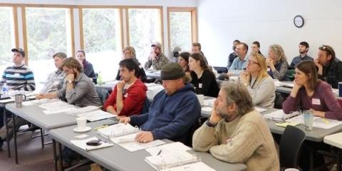 A photo of people sitting in a room at rows of tables facing the front of the room while listening to a presentation.